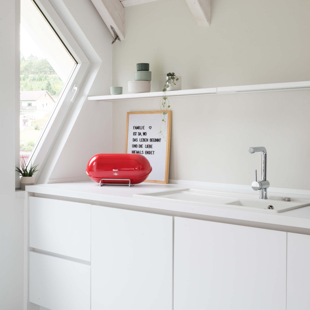 A modern white kitchen corner with a countertop, sink, open shelving, and a bright red countertop wesco breadboy bread bin near a window.