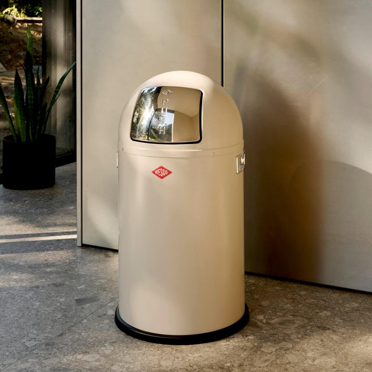 A cream coloured dome top waste bin with chrome push flap and handles. Black base ring placed on a stone floor and light grey kitchen cabinets.