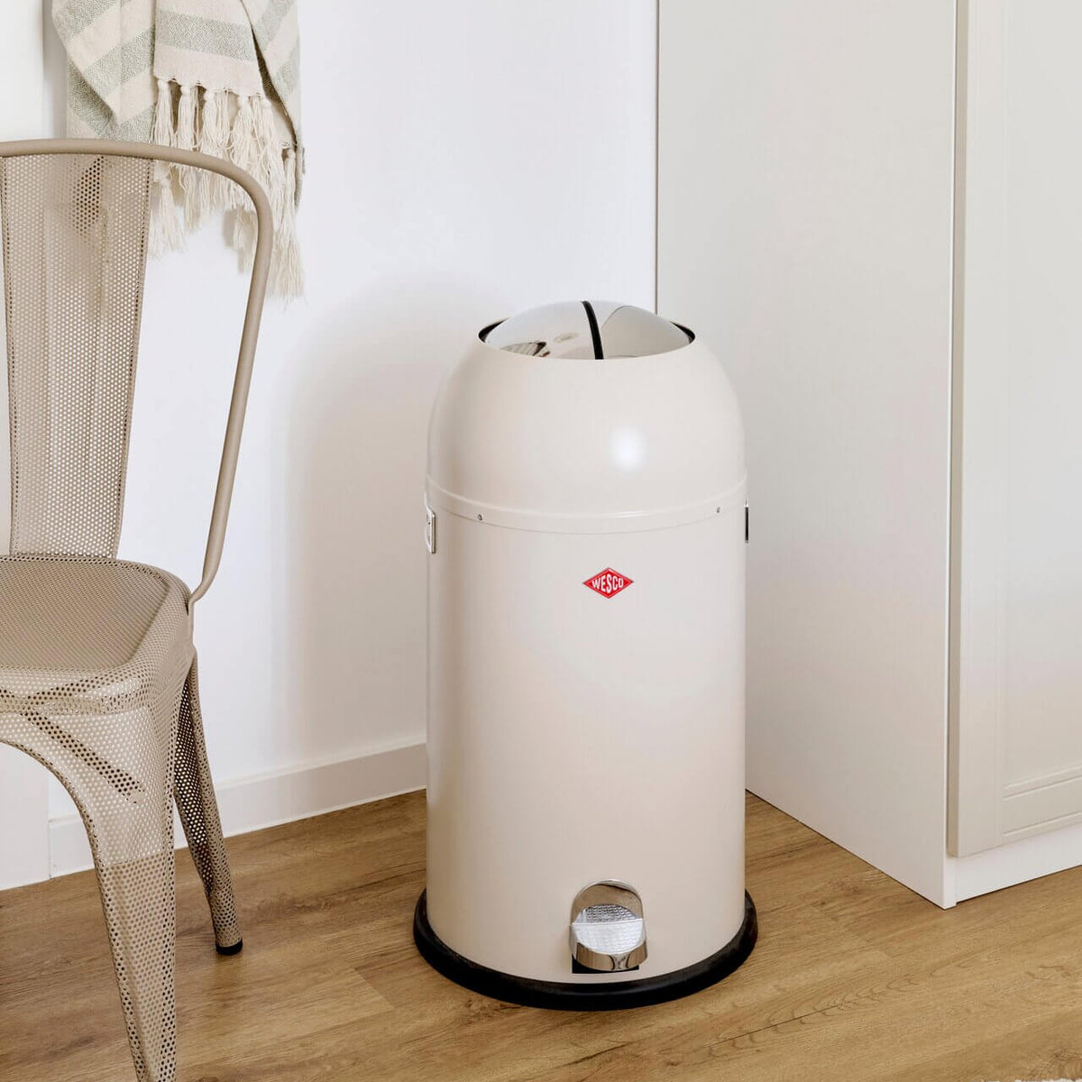 A cream-colored cylindrical pedal waste bin with a black base and a small red logo, placed on a light wood floor in a minimalist room next to a metal chair.