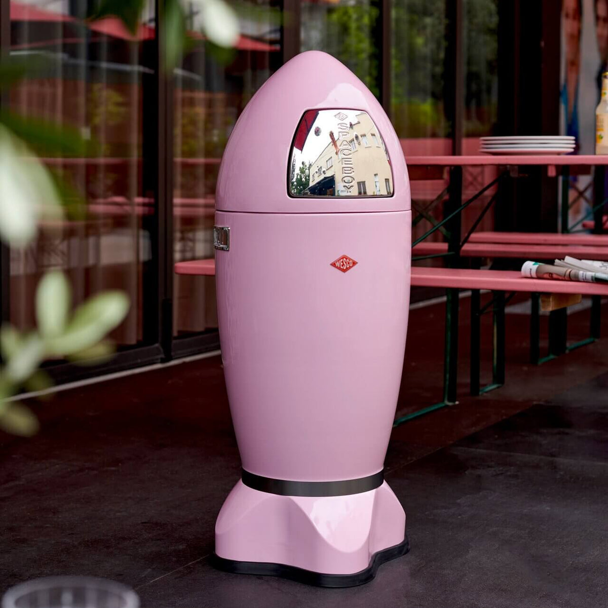 A pink push flap rocket-shaped trash, set on a dark flooring in front of a restaurant with pink tables and glass windows.