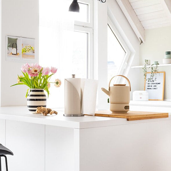 Wesco Kitchen roll holder in almond on a white kitchen with a kettle in cream and a white a black vase with pink and yellow flowers.