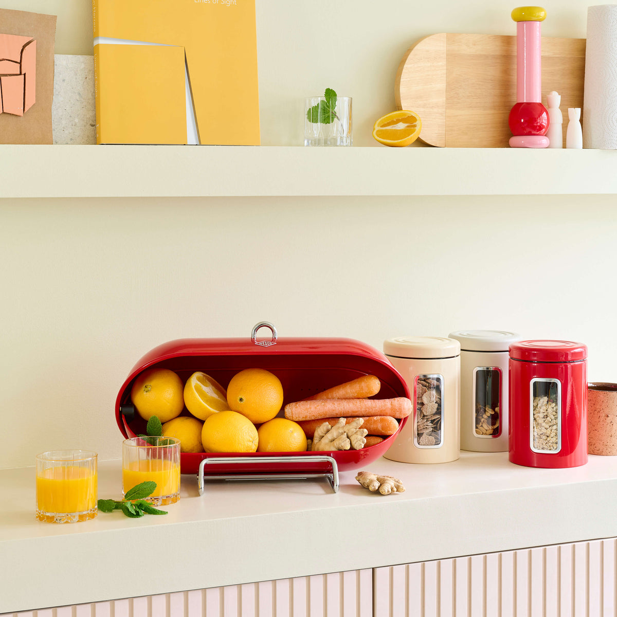 A red wesco breadboy metal bread bin filled with lemons, oranges, and carrots on a white kitchen counter, styled with matching canisters, glasses of juice, and a yellow book on white shelves above.