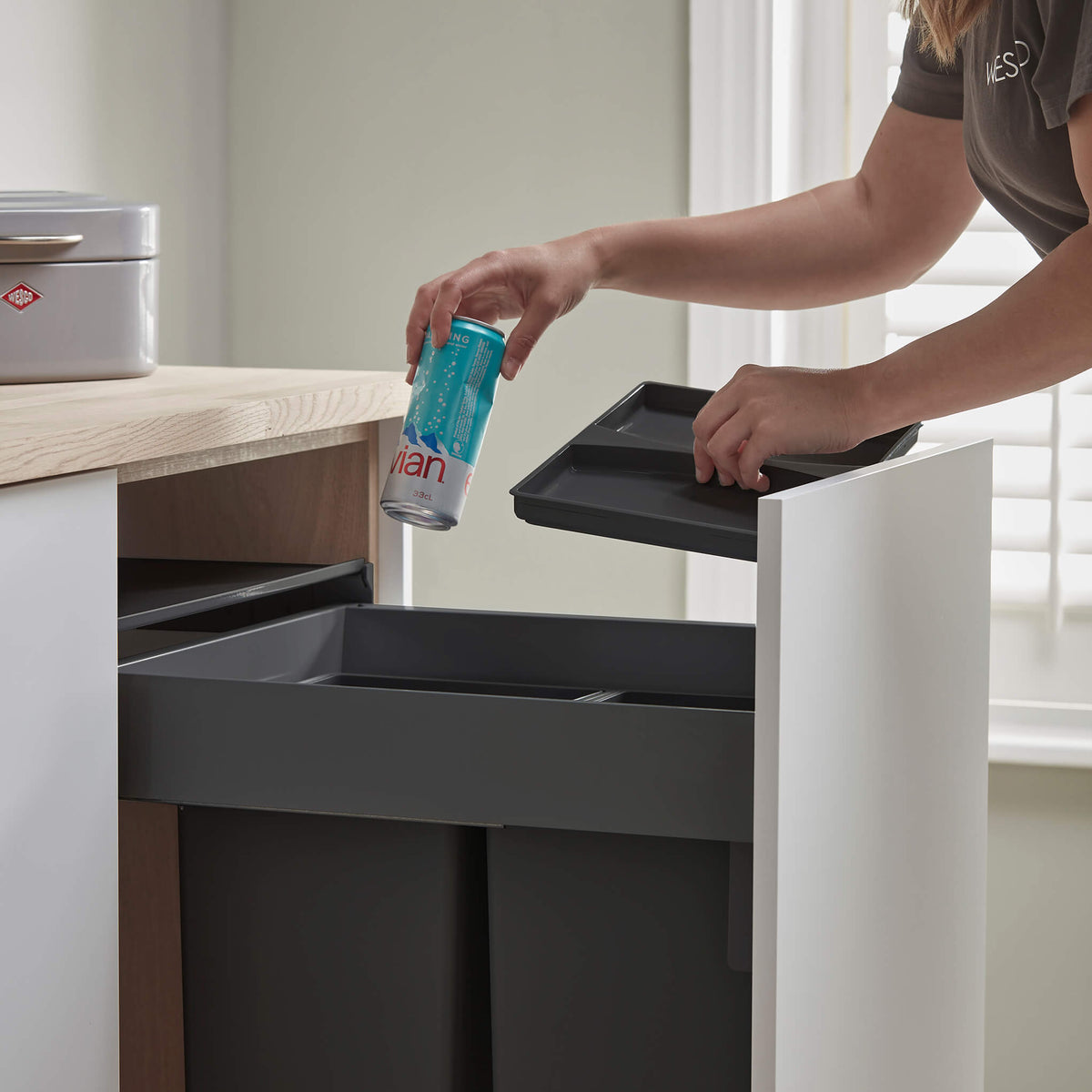 Person disposing of a can into a trash bin with a neutral background