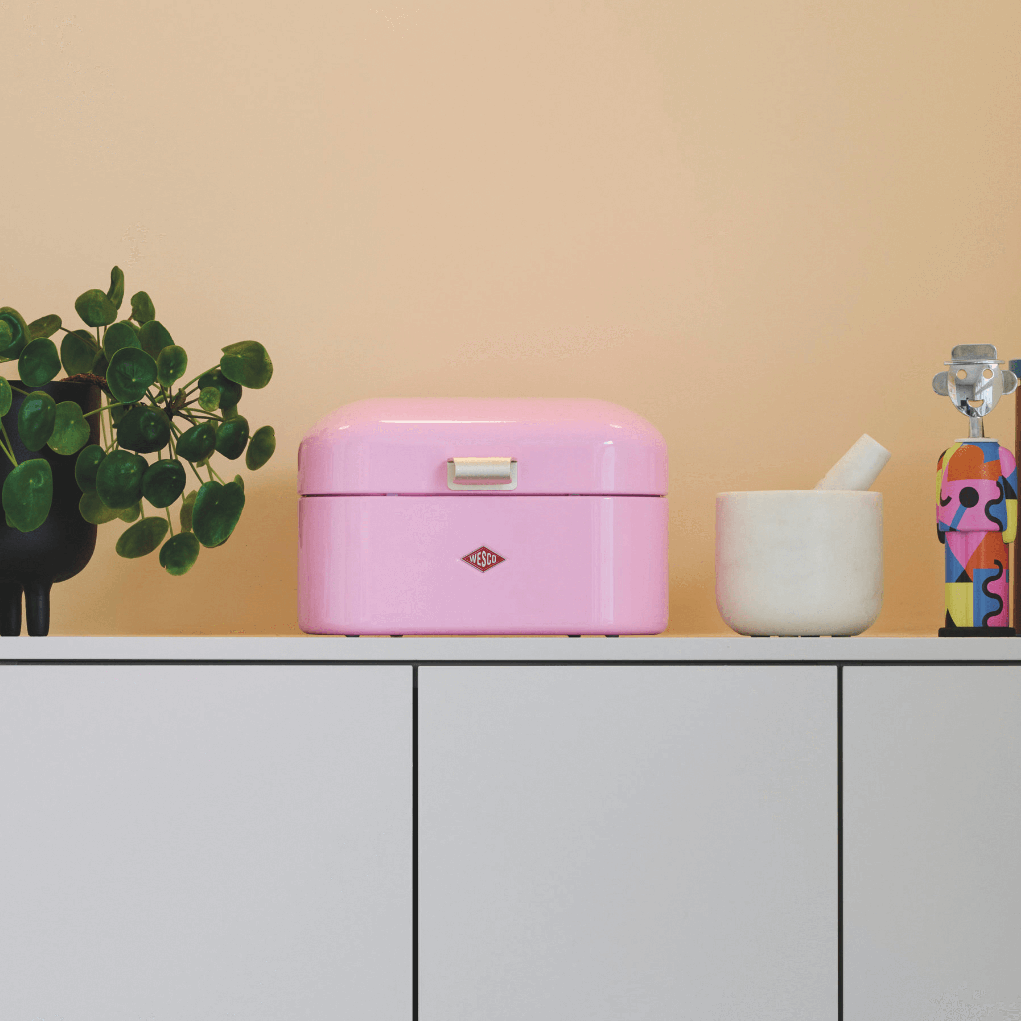 Pink bread bin on a white cabinet with a plant and decorative items in the background