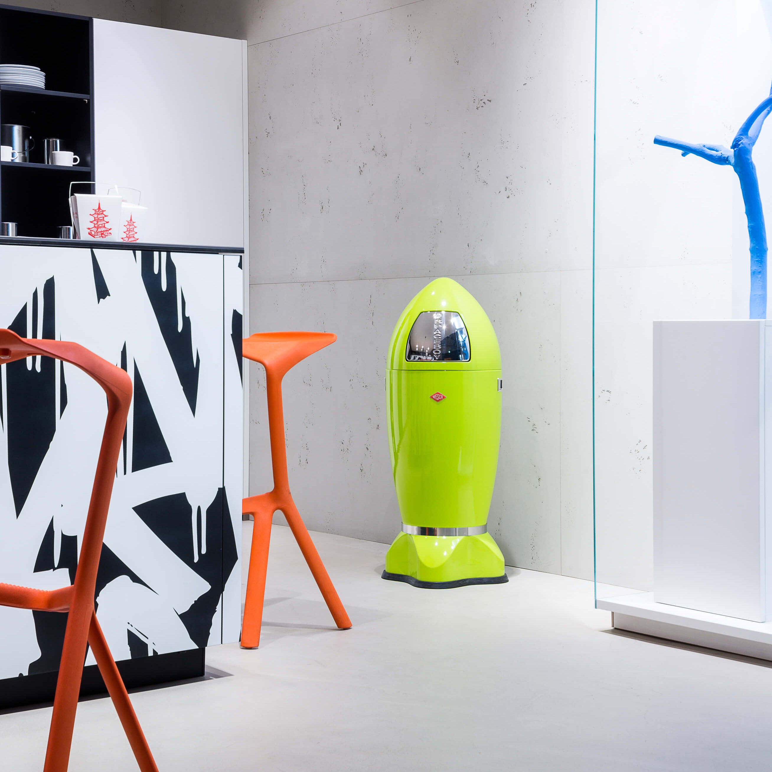 Modern interior with a lime green rocket shaped Spaceboy XL push flap waste bin device, orange chair, and black and white abstract art.