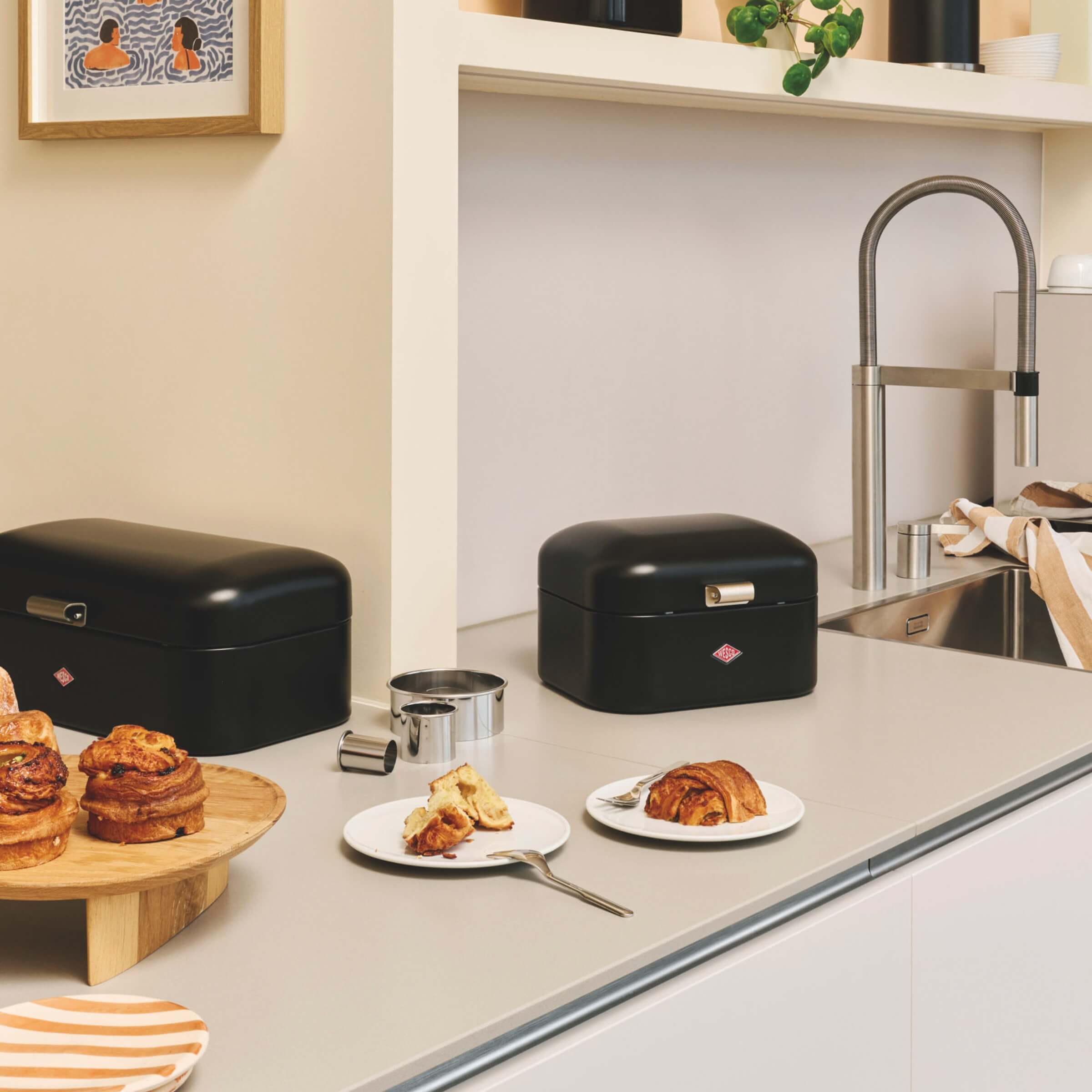 Black Wesco Grandy bread bins on a kitchen counter with pastries and plates.
