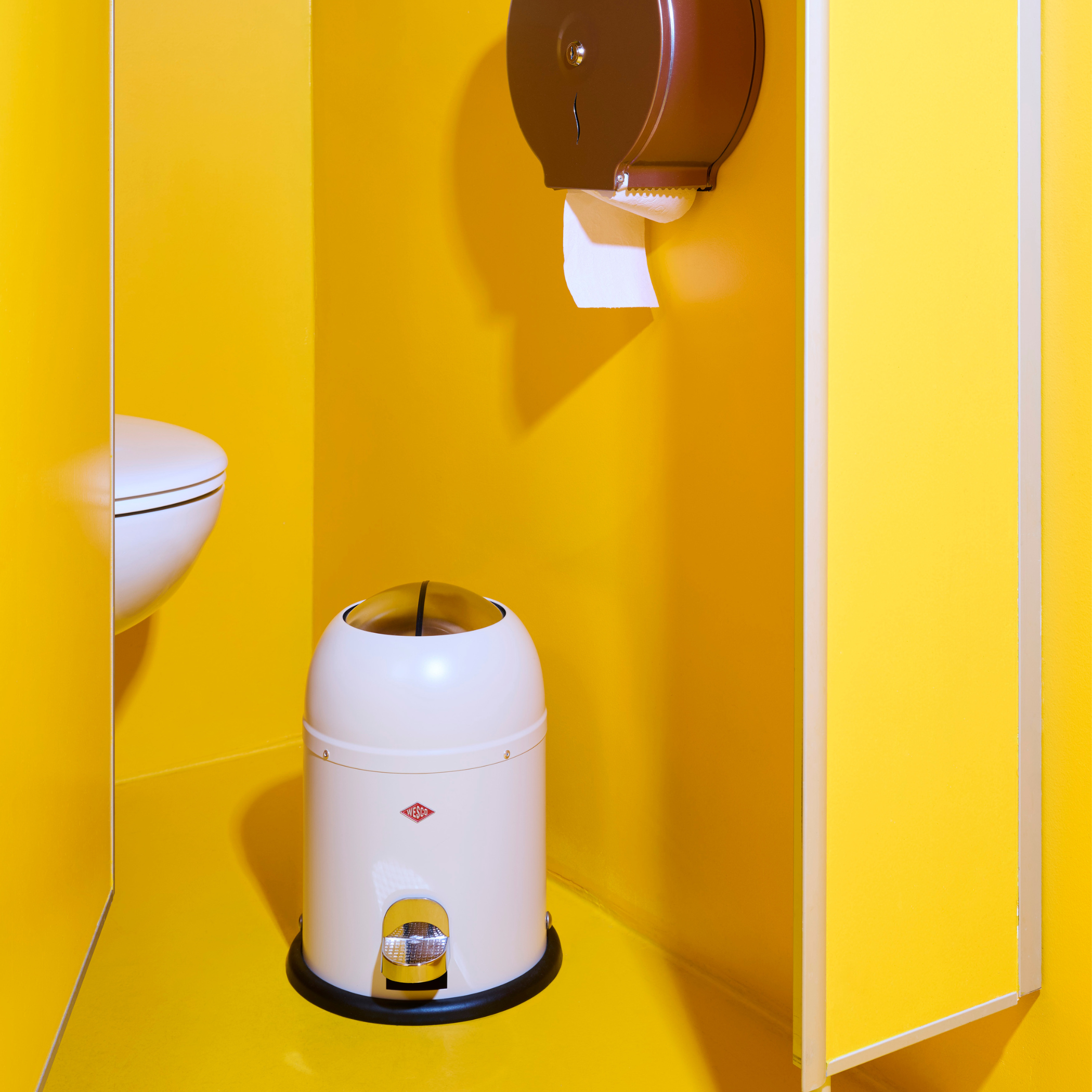 Wesco Matt White Mini Master Waste Bin with black base against a yellow wall with a toilet partially visible.