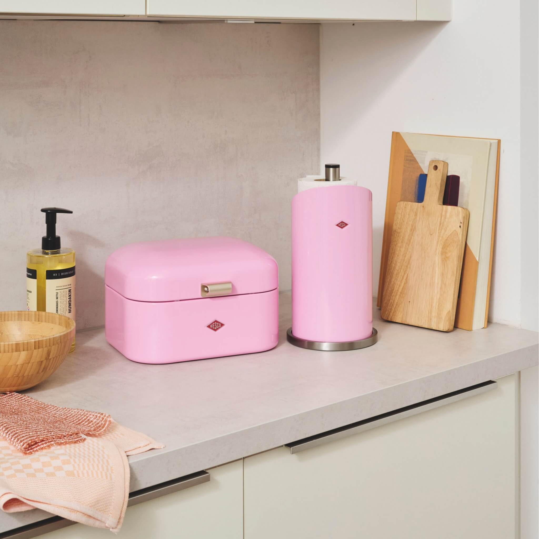 Wesco paper Roll holder in pink on a grey kitchen worktop along with the matching pink bread bin and wooden chopping boards