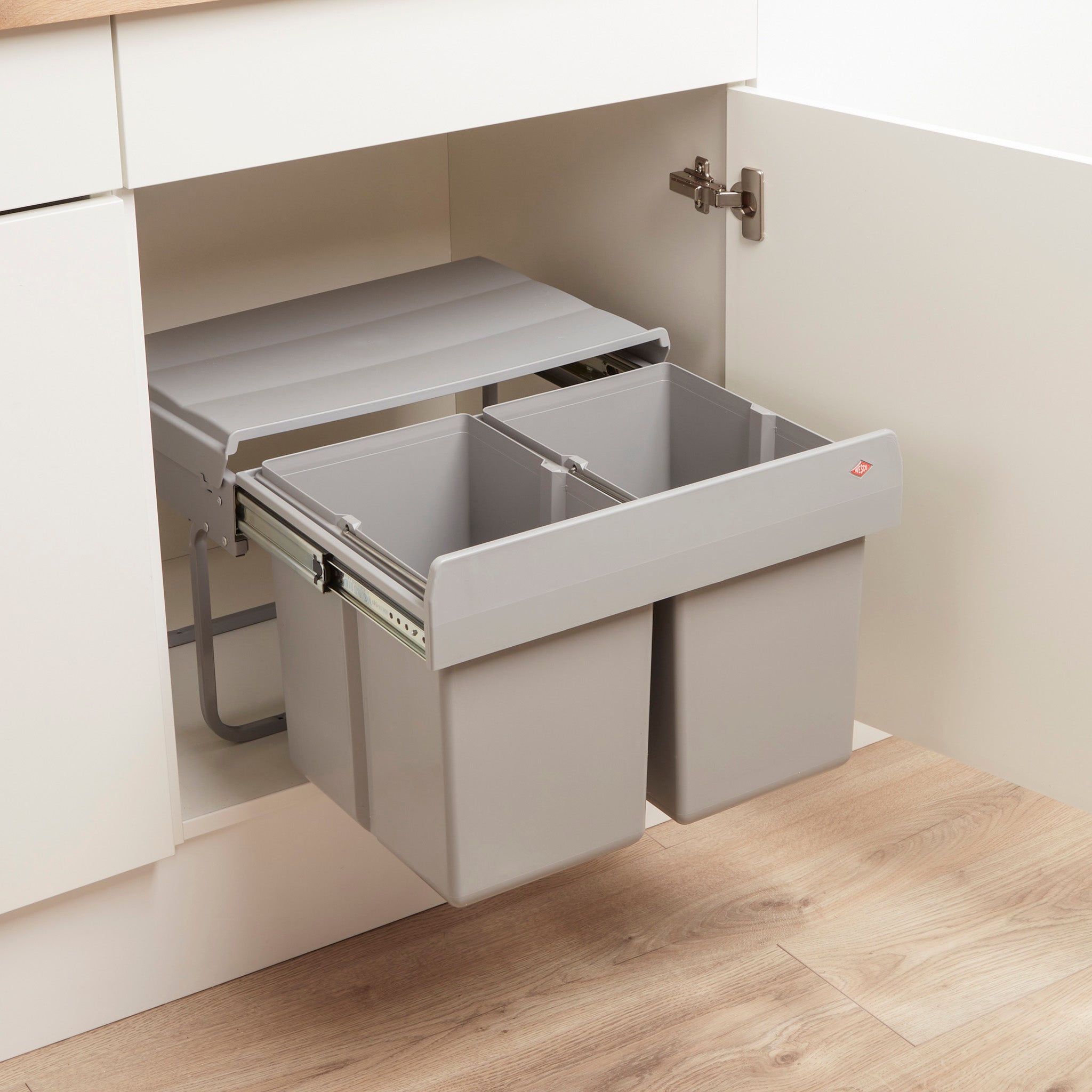 Wesco Shorty manual waste bin with two compartments, installed beneath a kitchen sink inside a cabinet with a hinged door.