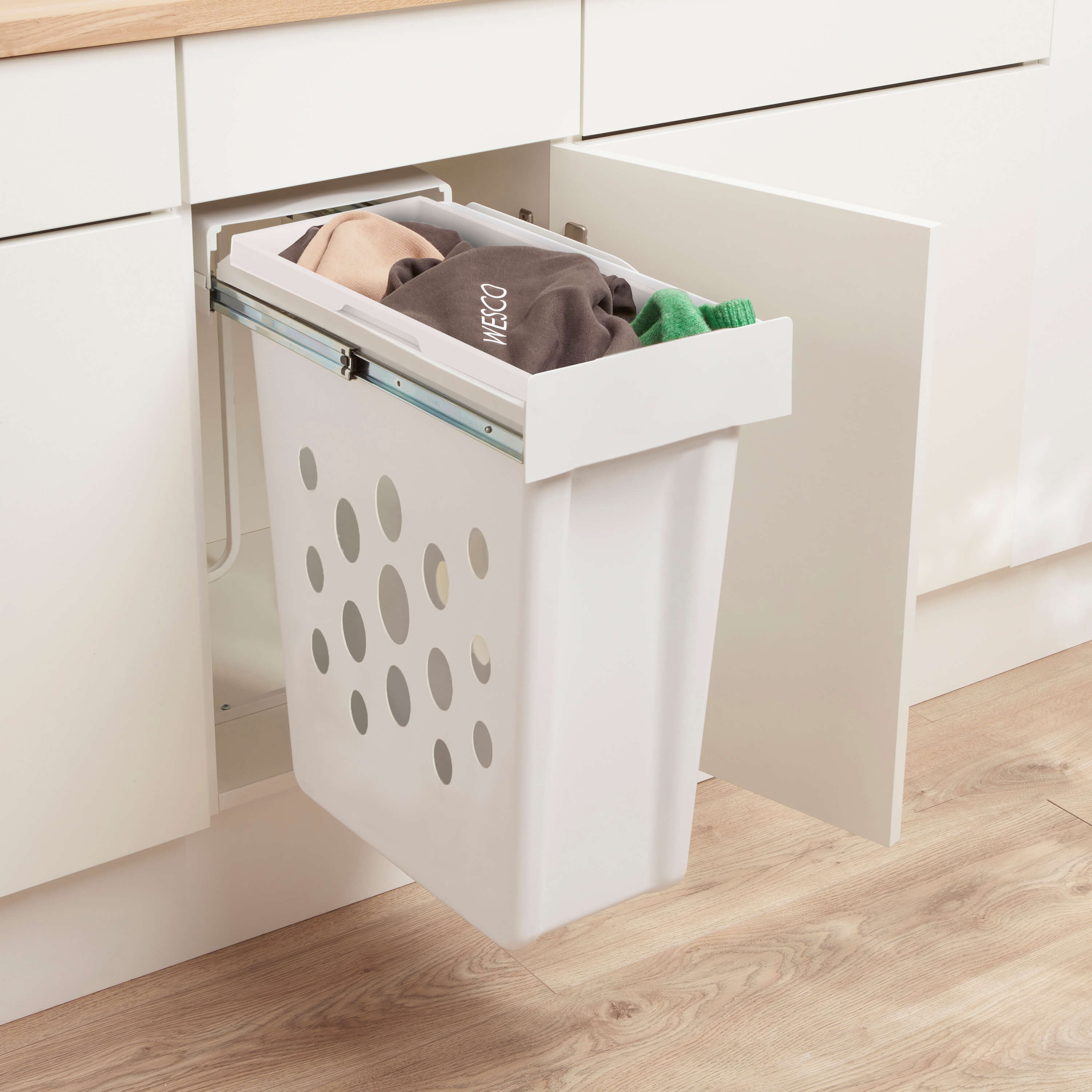 Wesco Laundry Boy with clothes in, a White laundry basket with perforated design on runners pulled out of a kitchen cabinet