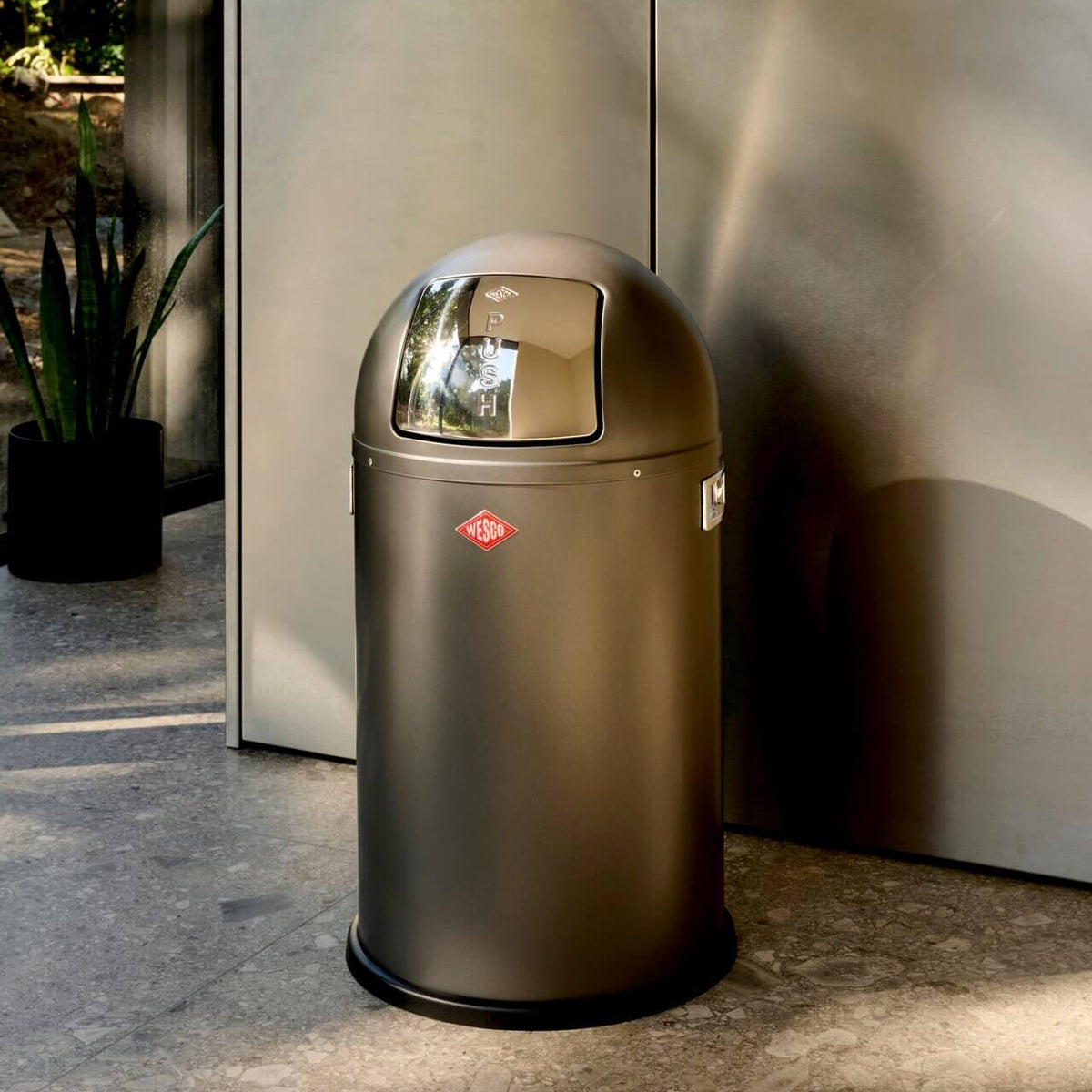 A modern dome waste bin with a brushed dark metallic finish, in grey kitchen with stone floor.