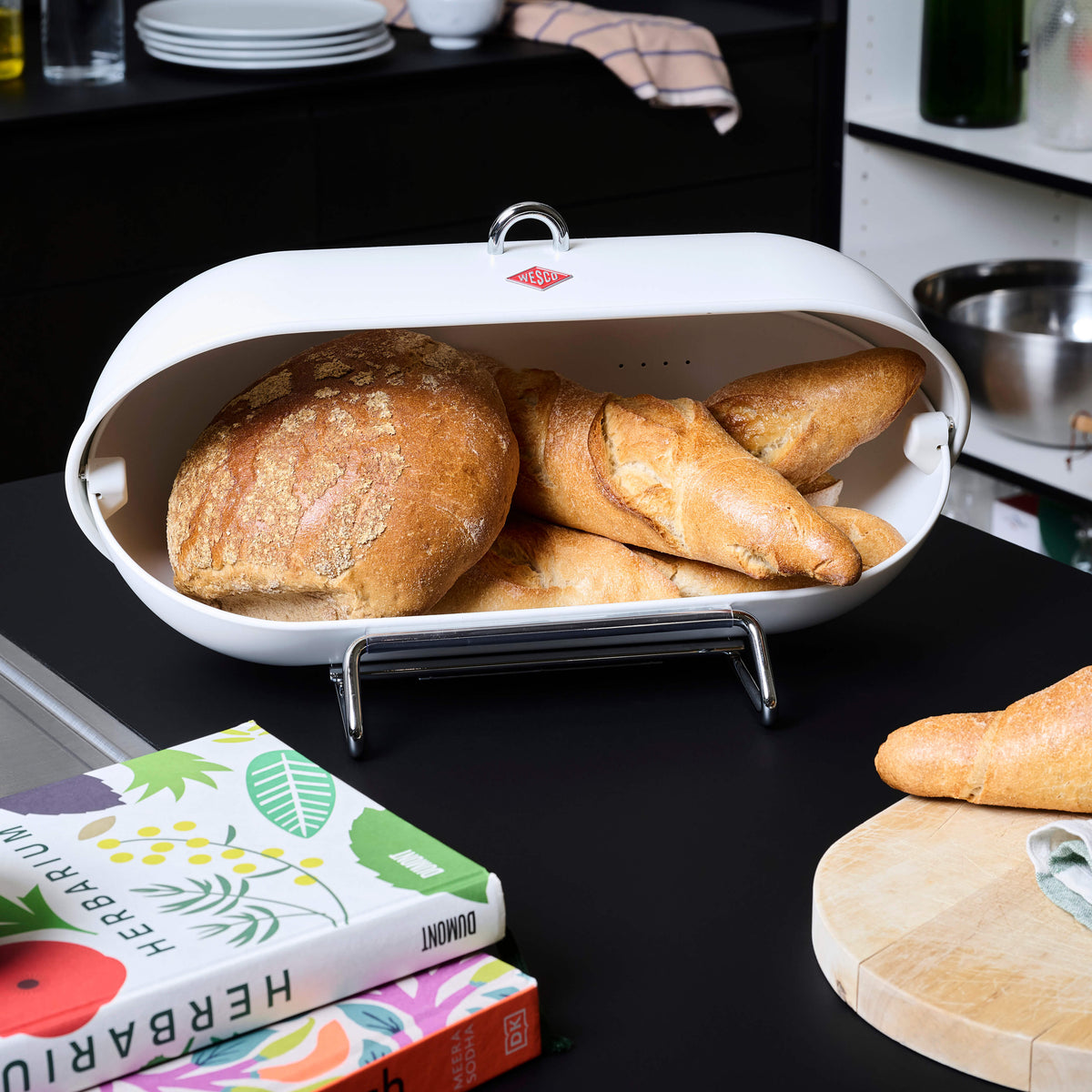 A white wesco breadboy bread bin with a metal handle and stand, filled with crusty bread loaves, set on a black countertop in a kitchen environment with bread, a cookbook, and other kitchen items around it.
