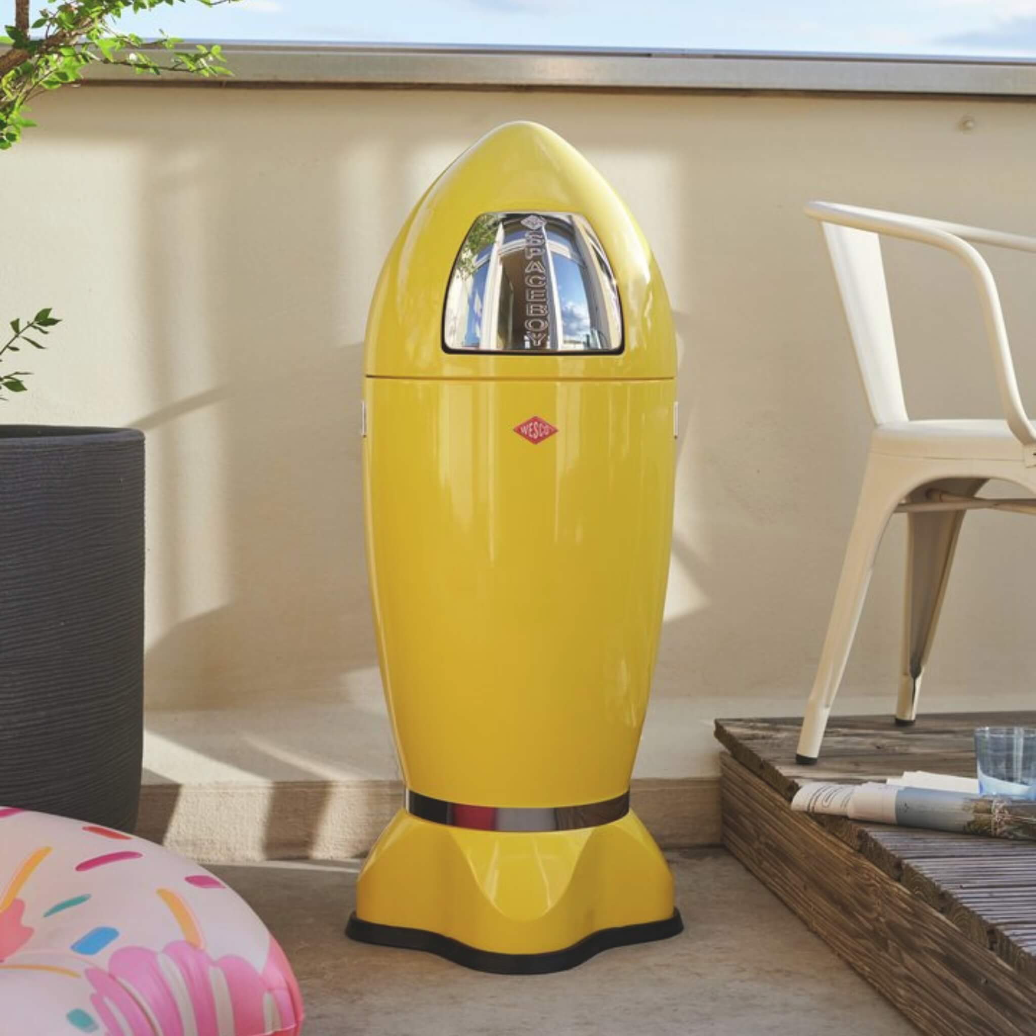 Wesco Spaceboy XL Yellow rocket-shaped Push Flap waste bin on a patio with a chair and table in the background.