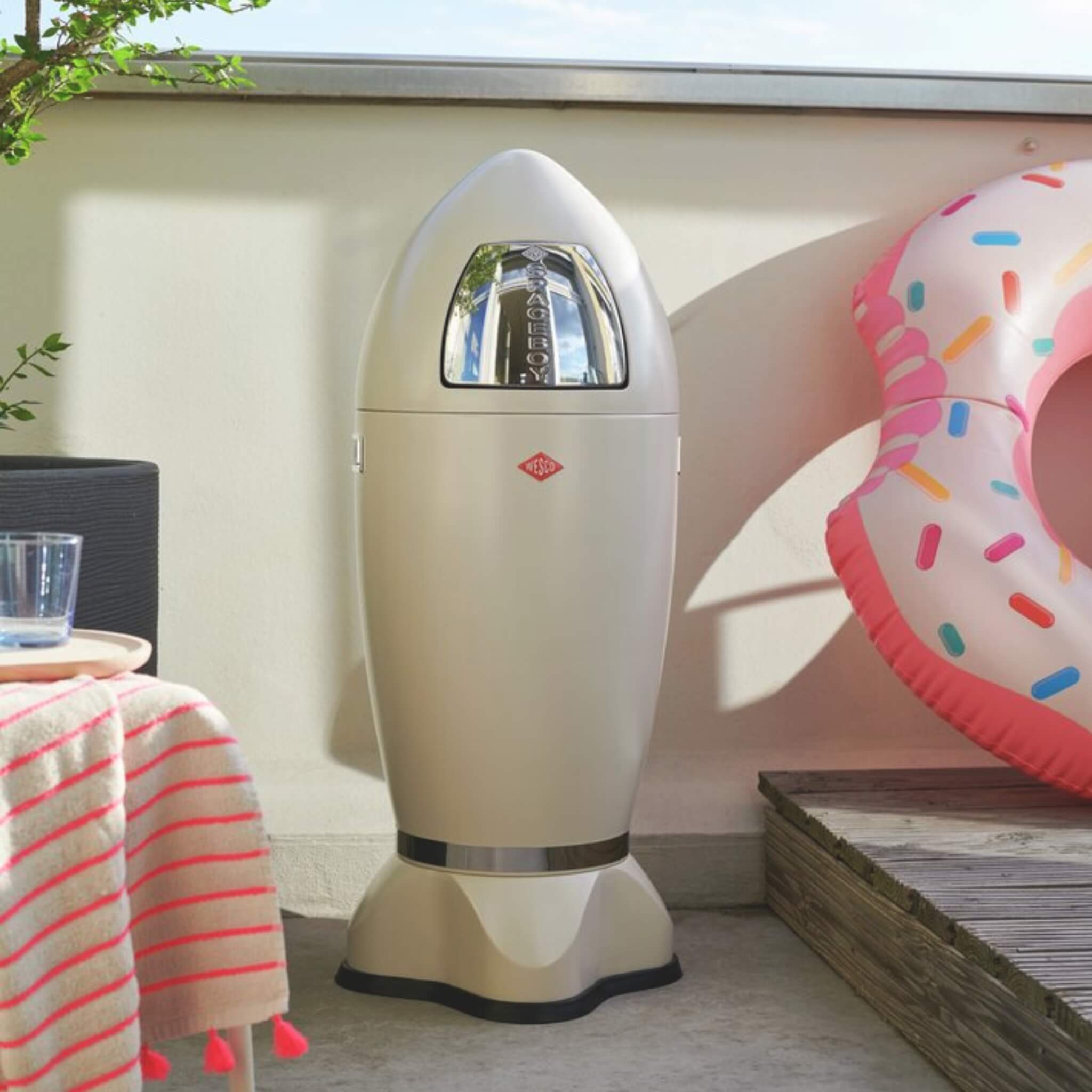 Wesco matt sand rocket-shaped push flap waste bin on a patio with a donut inflatable toy in the background.