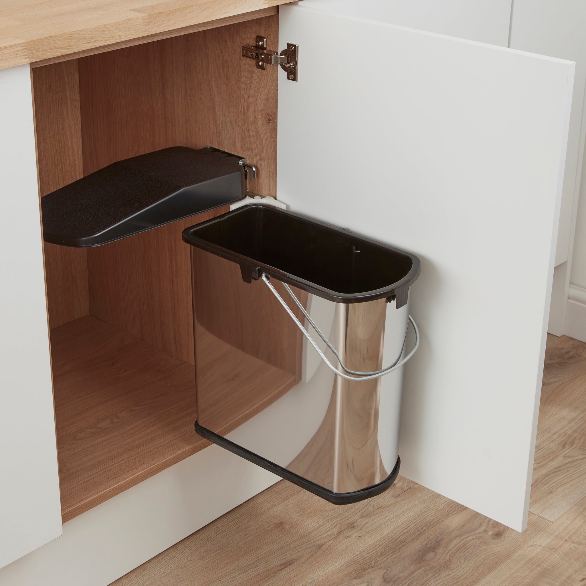 A slim-line stainless steel waste bin with a black lid, designed to fit under a kitchen sink within a cabinet, shown open with the bin swinging out.