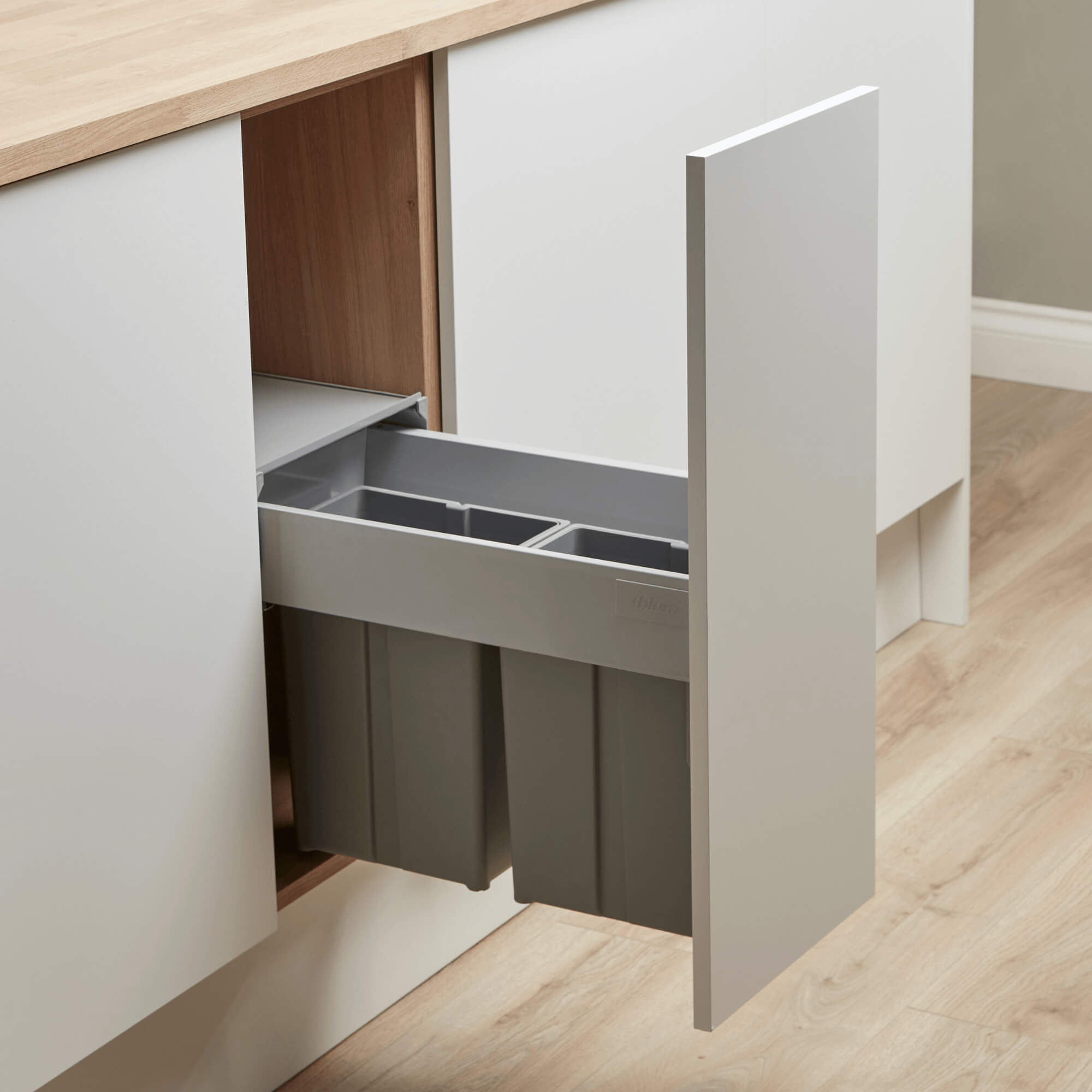An integrated recycling bin system installed under a kitchen counter, featuring a light grey steel frame and two plastic inner bins with sturdy handles, designed to fit into a 300mm wide cabinet.