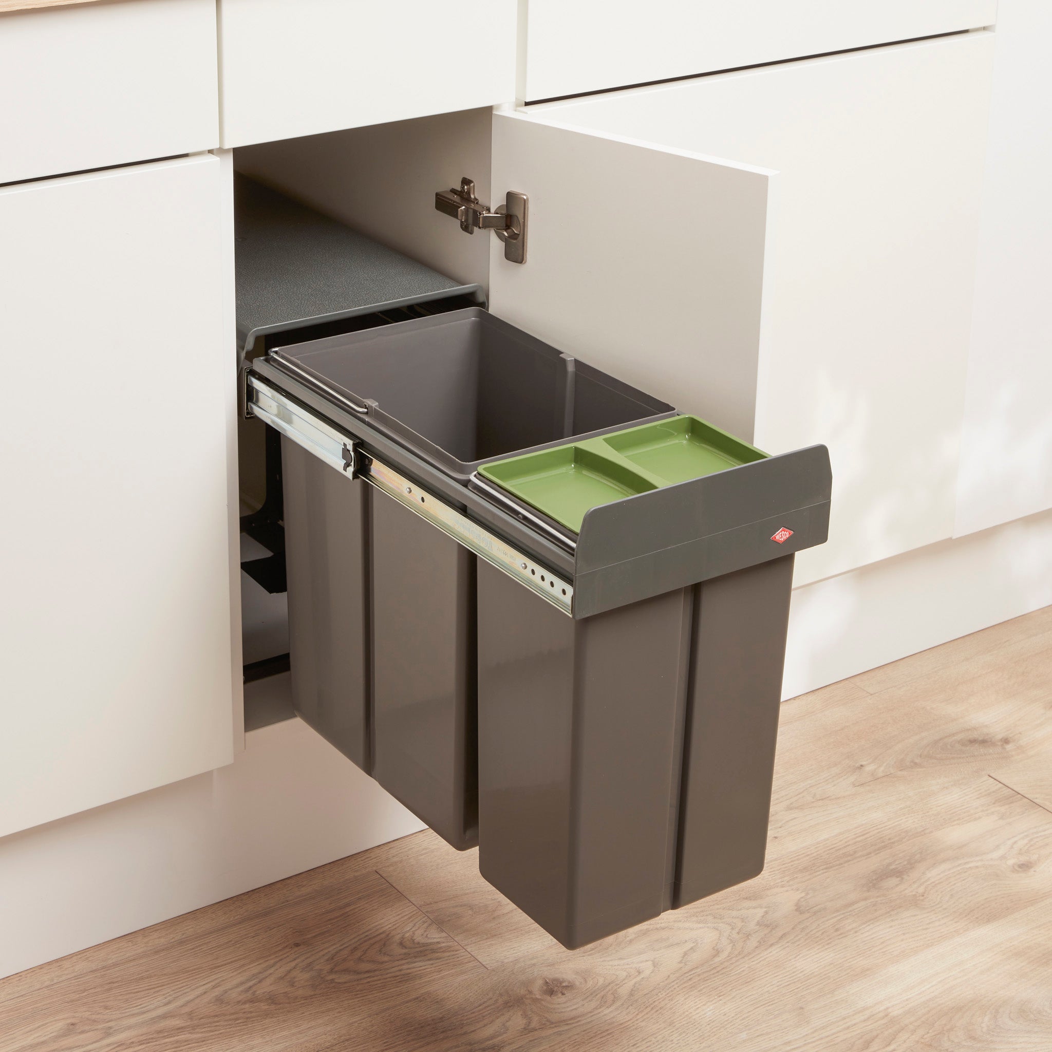 A Profiline Bio Double 30L waste bin with two compartments, Orion Grey, mounted to the base of a white cabinet with a green lid on the smaller compartment, visible through the open cabinet door.